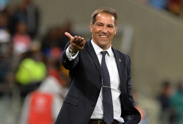 Man in a dark suit and tie smiles while extending his hand for a handshake on a sports field