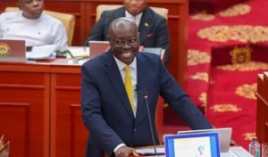 Smiling man in a navy suit and yellow tie speaks at a podium in a formal legislative chamber with red carpeted floors