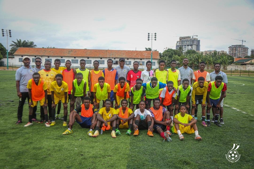 Group of young soccer players in bright bibs posing for a team photo on a grassy field, with coaches standing behind them and urban buildings in the background.