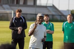 Coach in beige shirt and flat cap speaks to a small group of players on a sunny football field during training, holding glasses in hand and making a gesture.