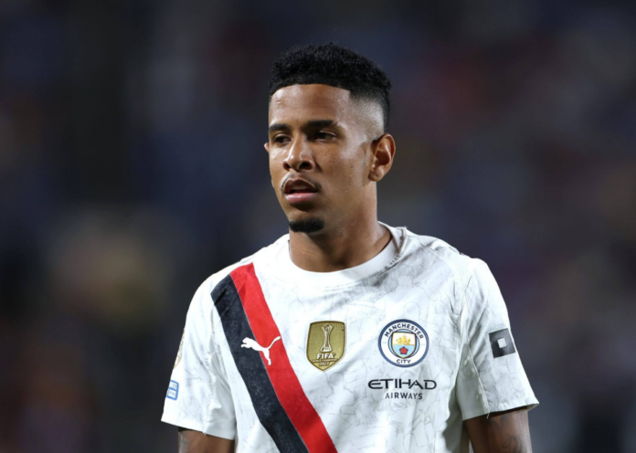 Footballer wearing a white Manchester City jersey on the field during a match with blurred stadium background.