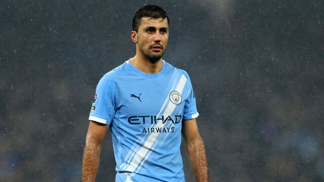 Soccer player wearing a light blue Manchester City jersey in the rain with the club crest visible on the chest.