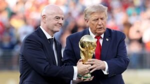 Two men in suits present a gold World Cup trophy on a stadium field, with a blurred crowd in the background.