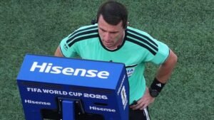 Soccer referee in a turquoise jersey leaning toward a blue FIFA World Cup 2026 monitor labeled Hisense on a grassy field.