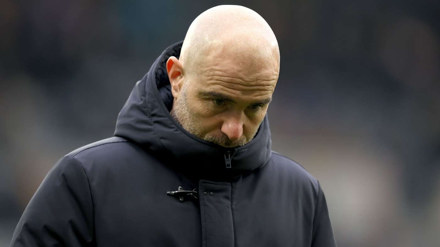 Bald man in a dark hooded jacket looking downward outdoors, with a blurred background in a sports setting.
