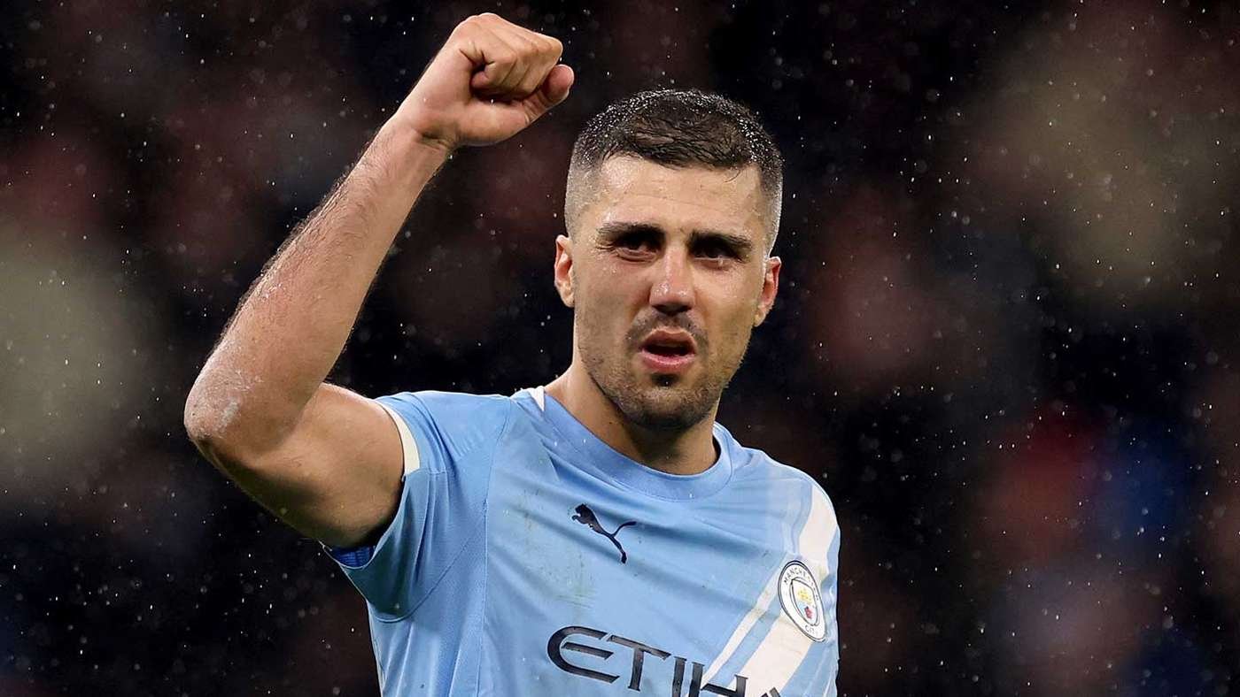 Manchester City footballer in a light-blue kit raises a clenched fist in celebration on a rain-soaked pitch.