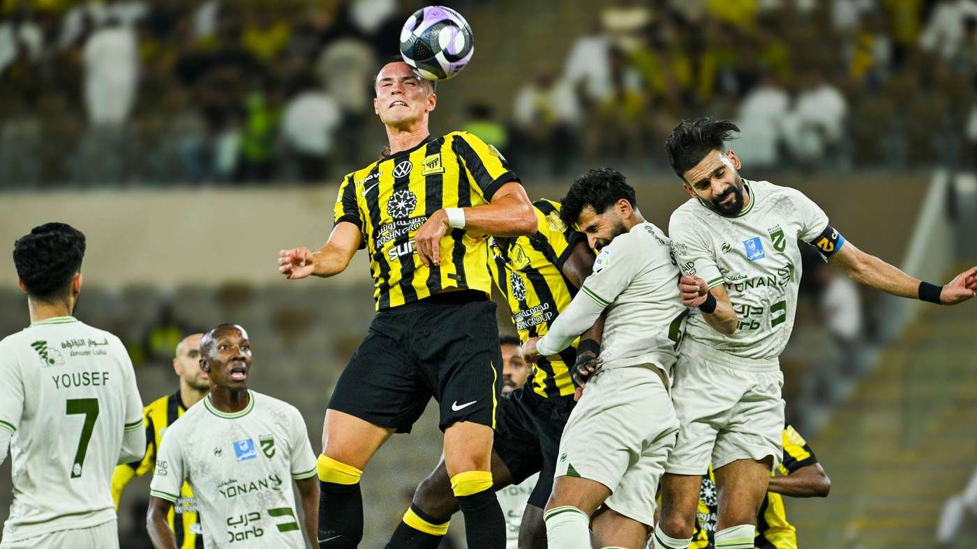 Soccer players in yellow-and-black and white kits contest a header for the ball in a packed stadium.