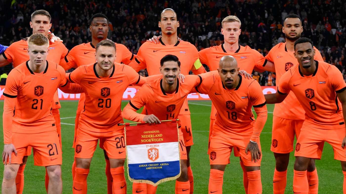 Dutch national football team players pose for a group photo in orange kits on a football pitch, holding a banner that reads the Netherlands vs Norway event.