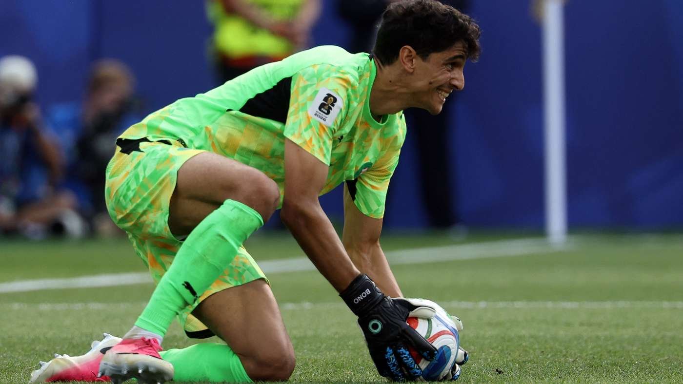 Goalkeeper in neon green kit kneels on the field, grabbing a soccer ball during a game action.