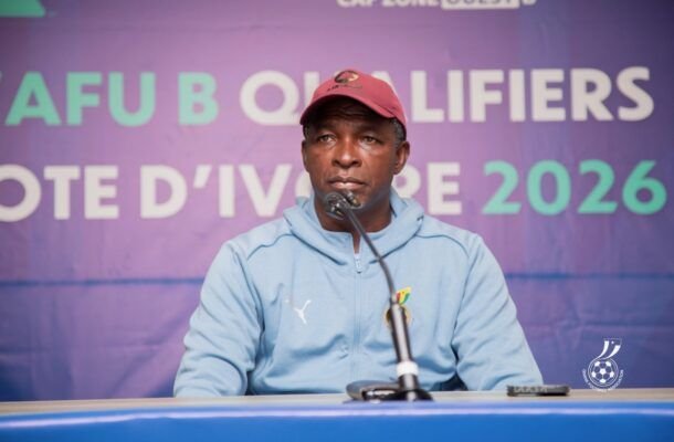 Man wearing a red cap and light blue hoodie sits at a microphone during a press conference with a purple AFU qualifiers banner behind