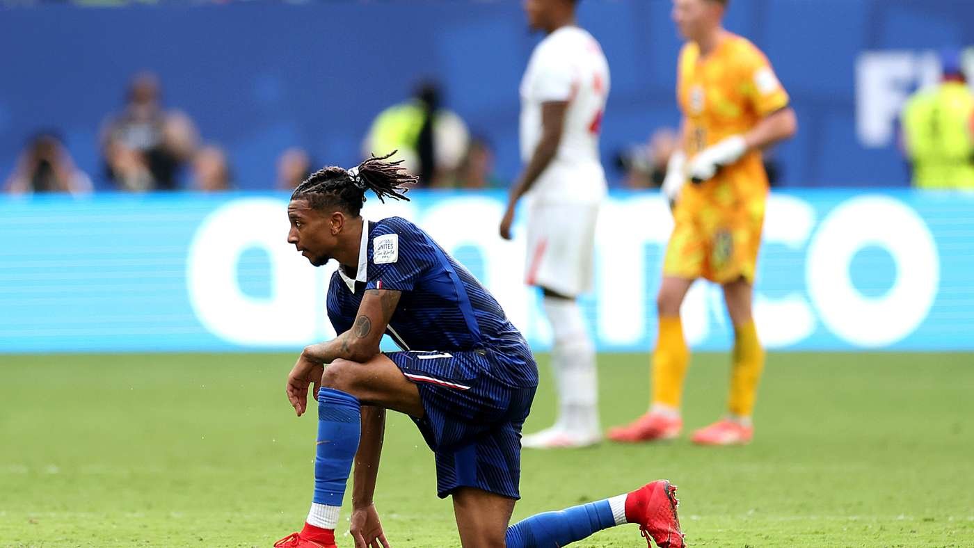 Soccer player in a blue uniform kneels on the green field during a match with two blurred players in white and yellow behind him
