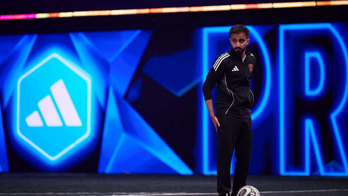 Man in black Adidas tracksuit standing on a soccer pitch with a ball at his feet blue neon background behind him