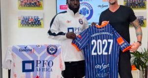 Two men pose with a blue striped Yamoussoukro FC 2027 jersey flanked by sponsor shirts and team photos on the wall behind them