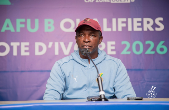 Man in a light blue hoodie and red cap sits at a press conference table speaking into a microphone with a purple banner reading CAF qualifiers behind him