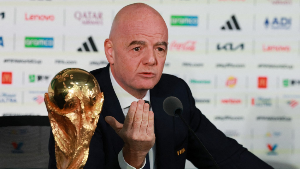Man in a suit at a press conference with a large golden FIFA World Cup trophy beside him on a table gesturing with his hand