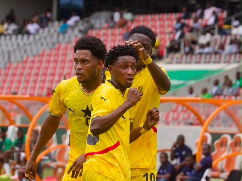 Three soccer players in yellow uniforms celebrate on the field near the sideline dugout raising their fists in victory