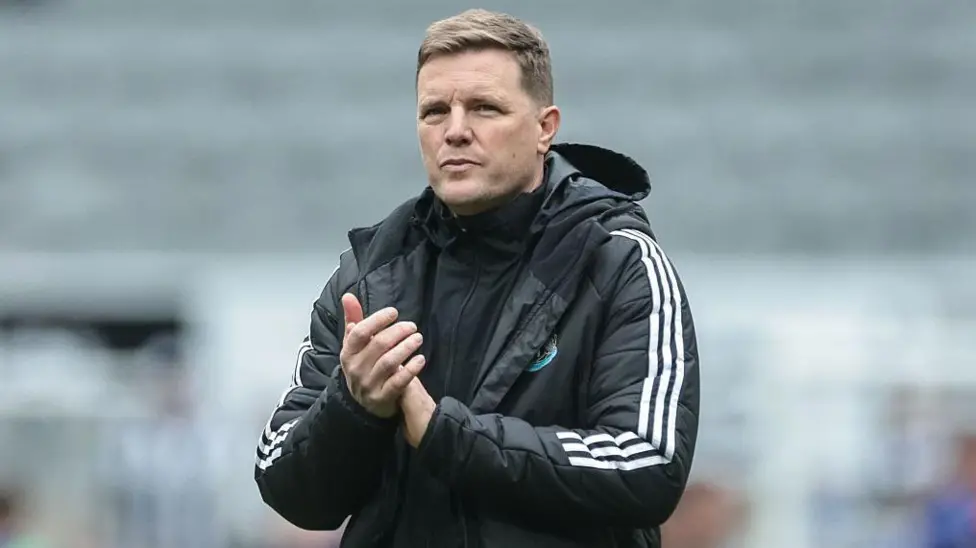 A man in a black sports jacket clapping standing outdoors on a field under overcast skies