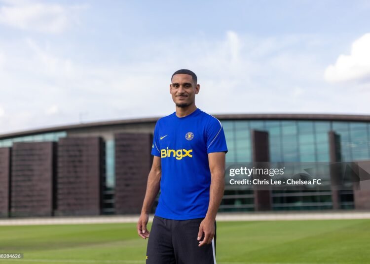 Male soccer player in a Chelsea blue training kit standing on a well kept training ground with a modern building in the background