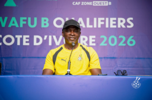 Man in a yellow athletic jersey speaks into a microphone at a blue podium during a press conference with a purple banner behind him reading QUALIFIERS and 2026