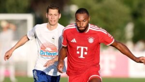 Two soccer players chase the ball during a match one in white with an orange sponsor the other in a red Bayern Munich kit on a sunny field momentarily contesting possession