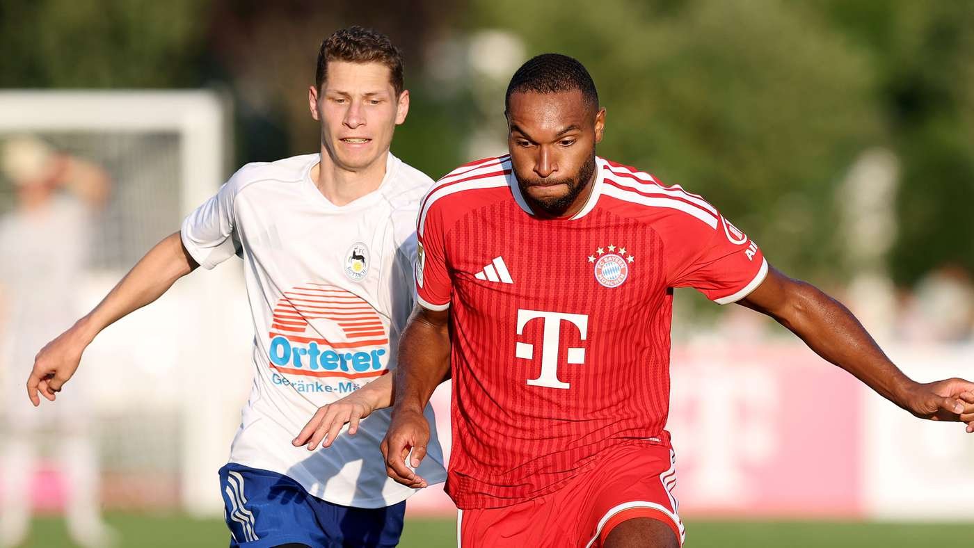 Two soccer players chase the ball during a match one in white with an orange sponsor the other in a red Bayern Munich kit on a sunny field momentarily contesting possession