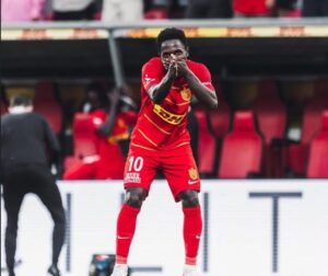 Soccer player in a red kit celebrating a goal cupping hands to mouth on the pitch with red seats behind