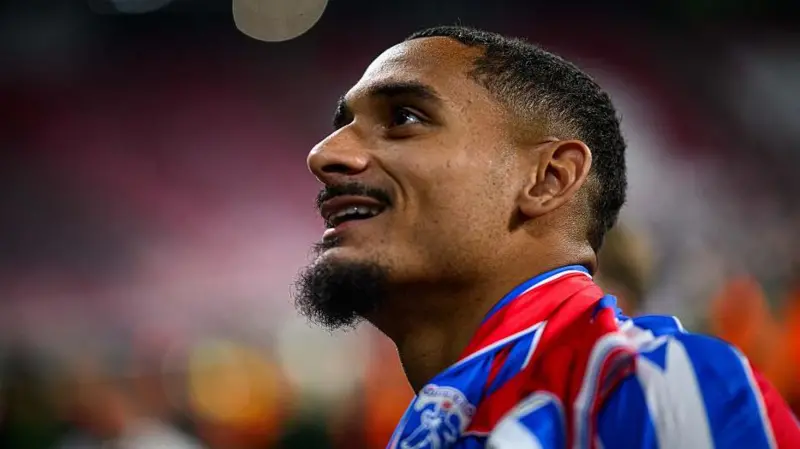 Soccer player in a red and blue striped jersey gazing upward at an approaching ball during a match