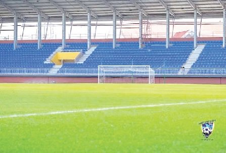 View of an empty soccer stadium with a green pitch white goal and blue seating stands in the background team crest visible in the bottom right corner