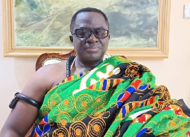 Man in a colorful traditional wrap and sunglasses sits in a chair indoors with a framed painting in the background