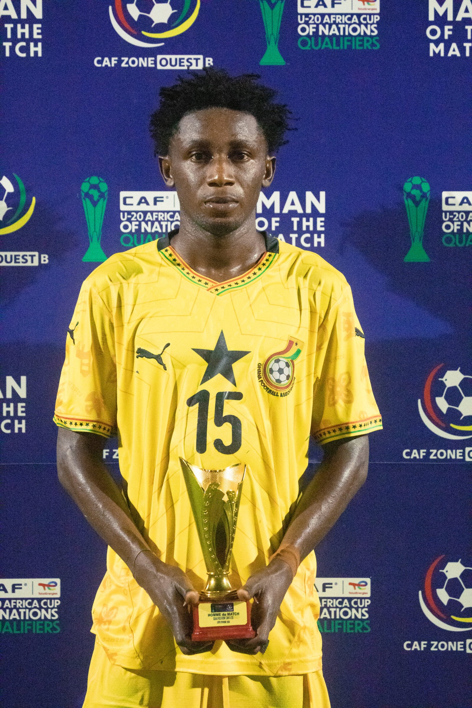 Young male soccer player in a yellow jersey holding a trophy standing in front of a blue backdrop with CAF Africa Cup logos