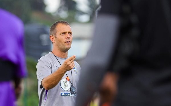 Coach or trainer in a gray shirt speaks and points during a practice on the field with players nearby in blurred foregrounds