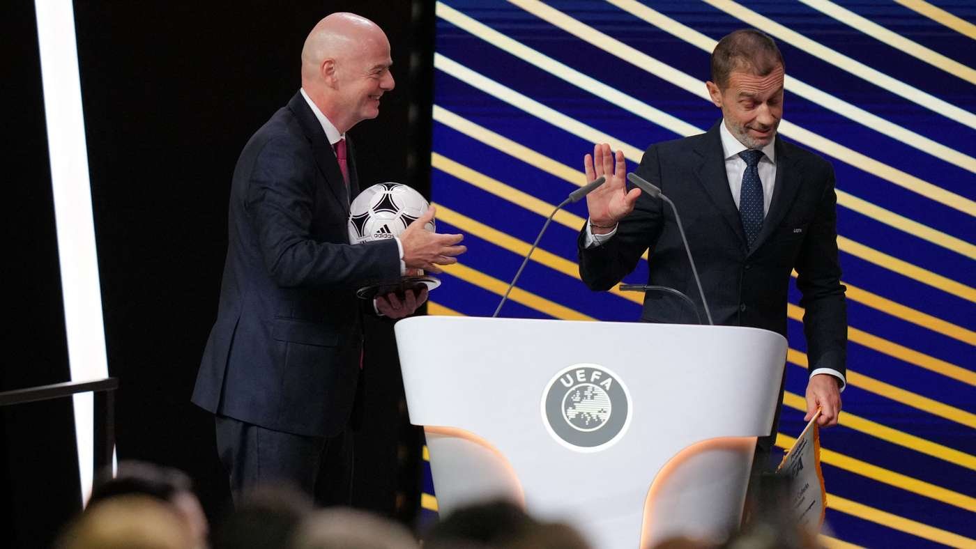 Two men in suits on a stage with a UEFA podium one holds a soccer ball and smiles while the other gestures farewell or speaks