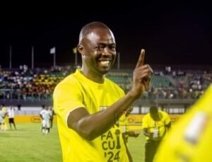 Smiling male soccer player in a yellow jersey on a lit stadium field at night raising one finger in celebration
