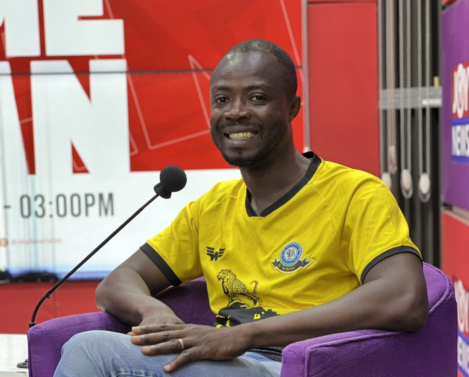 Man in a bright yellow shirt sits in a purple chair speaking into a handheld microphone during an interview or talk show The setting is a studio with red and purple signage in the background