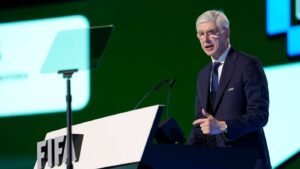 Older man in a dark suit speaks at a podium with the FIFA logo on stage microphones in front of him