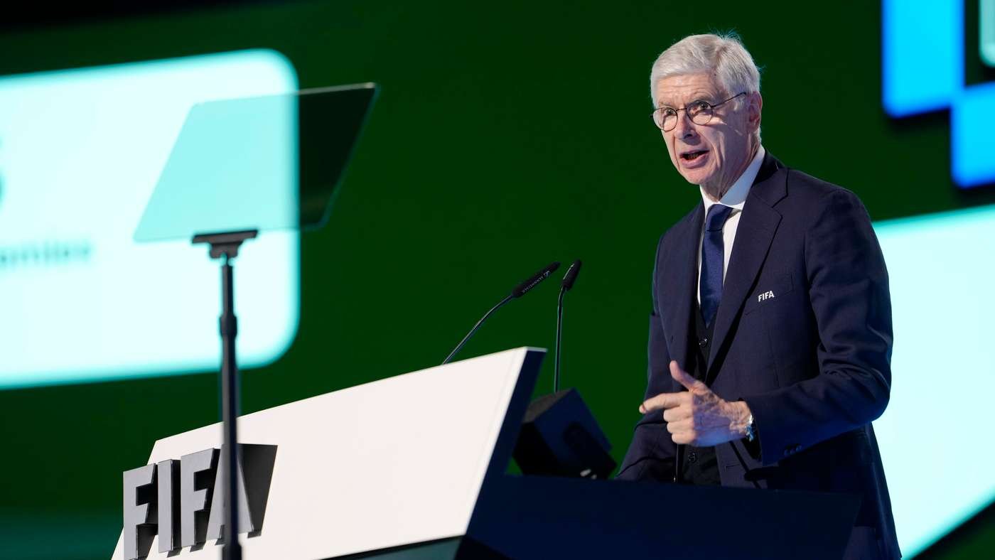 Older man in a dark suit speaks at a podium with the FIFA logo on stage microphones in front of him
