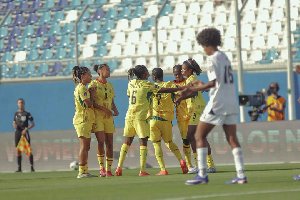 Female soccer players in yellow uniforms celebrate together on the green field as a player in white looks on nearby in the stadium
