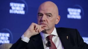 Bald man in a dark suit and red tie sits on stage hand resting on chin wearing a clip on microphone against a blue conference backdrop