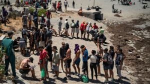Long queue of people on a sandy beach many with backpacks waiting near a jetty or barrier