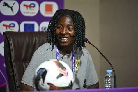 Smiling woman at a press conference table with a soccer ball in the foreground and a microphone nearby