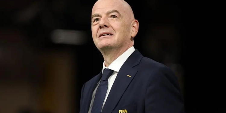 Bald man in a dark suit and navy tie with a FIFA pin looking upward as if speaking at an event