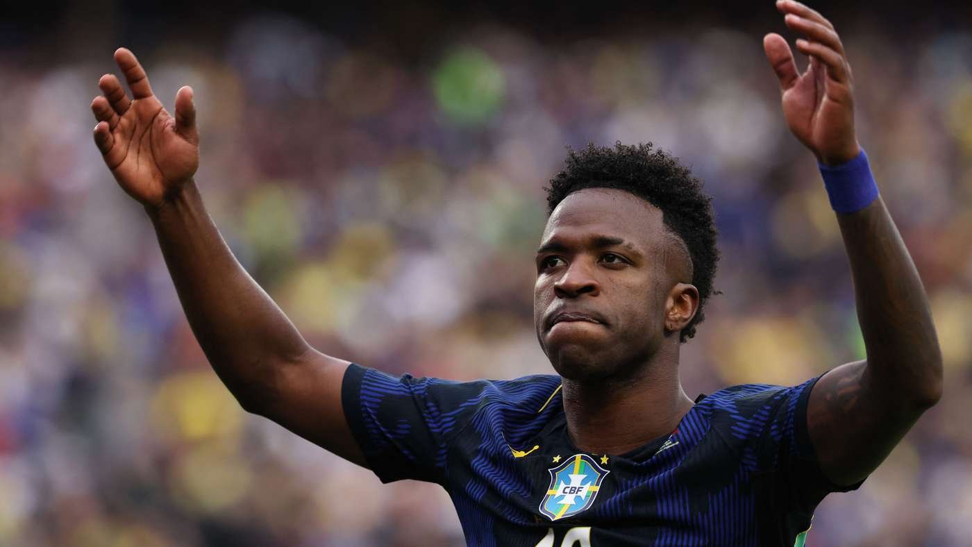 Brazilian footballer in blue Brazil kit raising arms in celebration on the pitch outside the stadium crowd