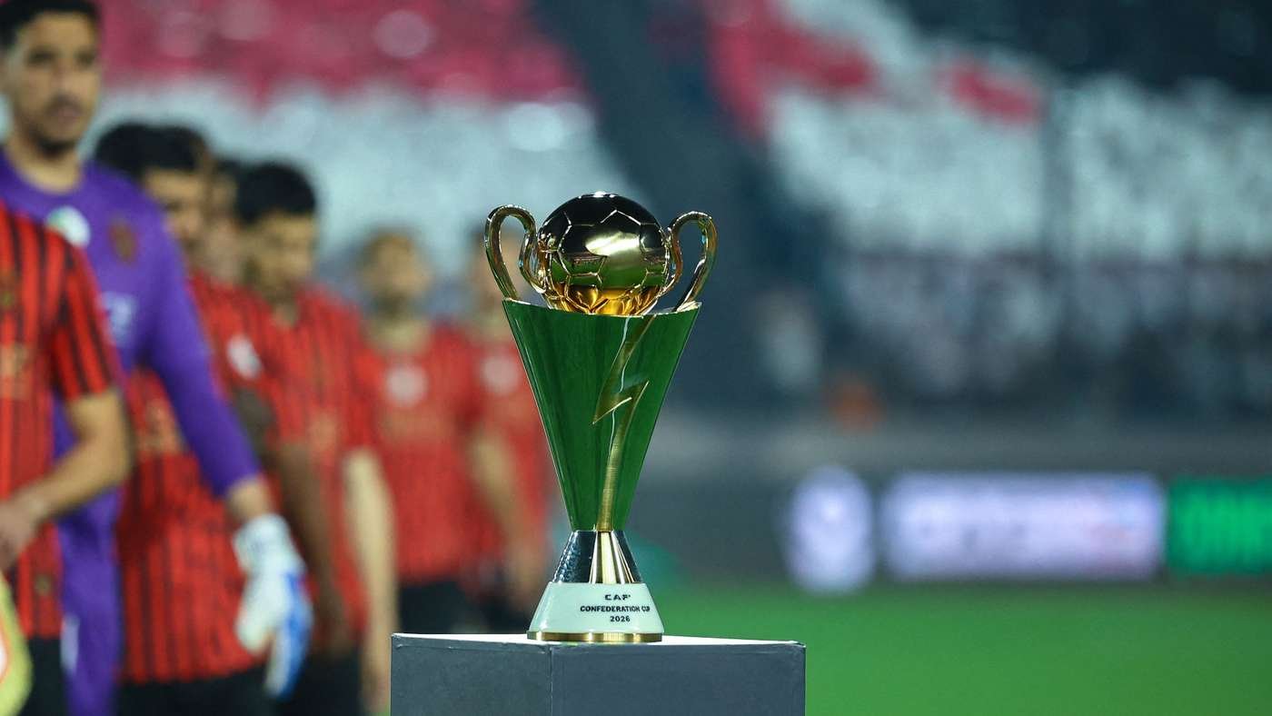Trophy on a pedestal in a stadium with a blurred line of players in red jerseys behind it