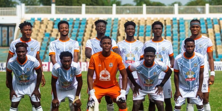 Professional soccer team poses for a group photo on a grass field players wear white kits with orange accents goalkeeper in orange stands behind them in a stadium