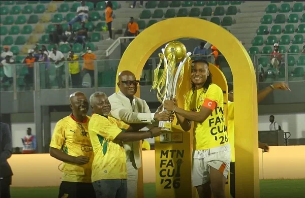 Football players in yellow celebrate on a pitch as a teammate accepts a trophy from officials under a large yellow arch centerpiece