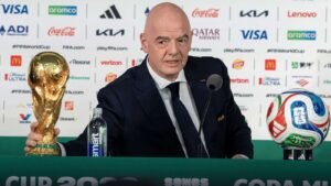 Bald man in a dark suit at a FIFA press conference holding the FIFA World Cup trophy a colorful ball sits on a stand to his right