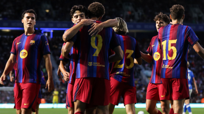 Barcelona football players in blue and red striped kits celebrate on the pitch embracing after a goal with teammates nearby and a stadium crowd in the background