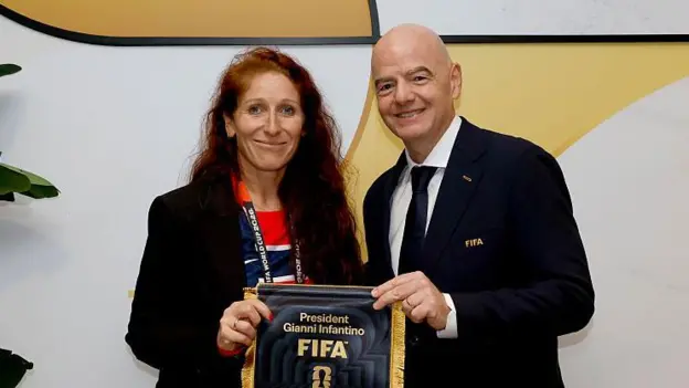 Two smiling officials in formal attire hold a dark commemorative plaque with FIFA branding posing for a photo against a light abstract backdrop