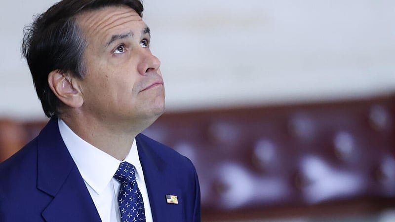 Man in a navy suit and tie looks upward with a thoughtful expression in an indoor setting