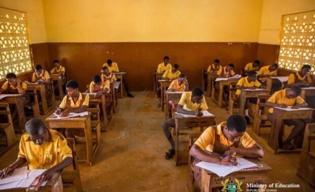 A classroom filled with students in yellow uniforms sitting at wooden desks writing on papers during an exam in a sunlit room